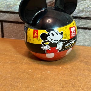 May include: A Mickey Mouse-shaped container with black ears and a black and yellow body. The container features a Mickey Mouse graphic and the text "90 Years of Magic". The bottom half is red with white polka dots.