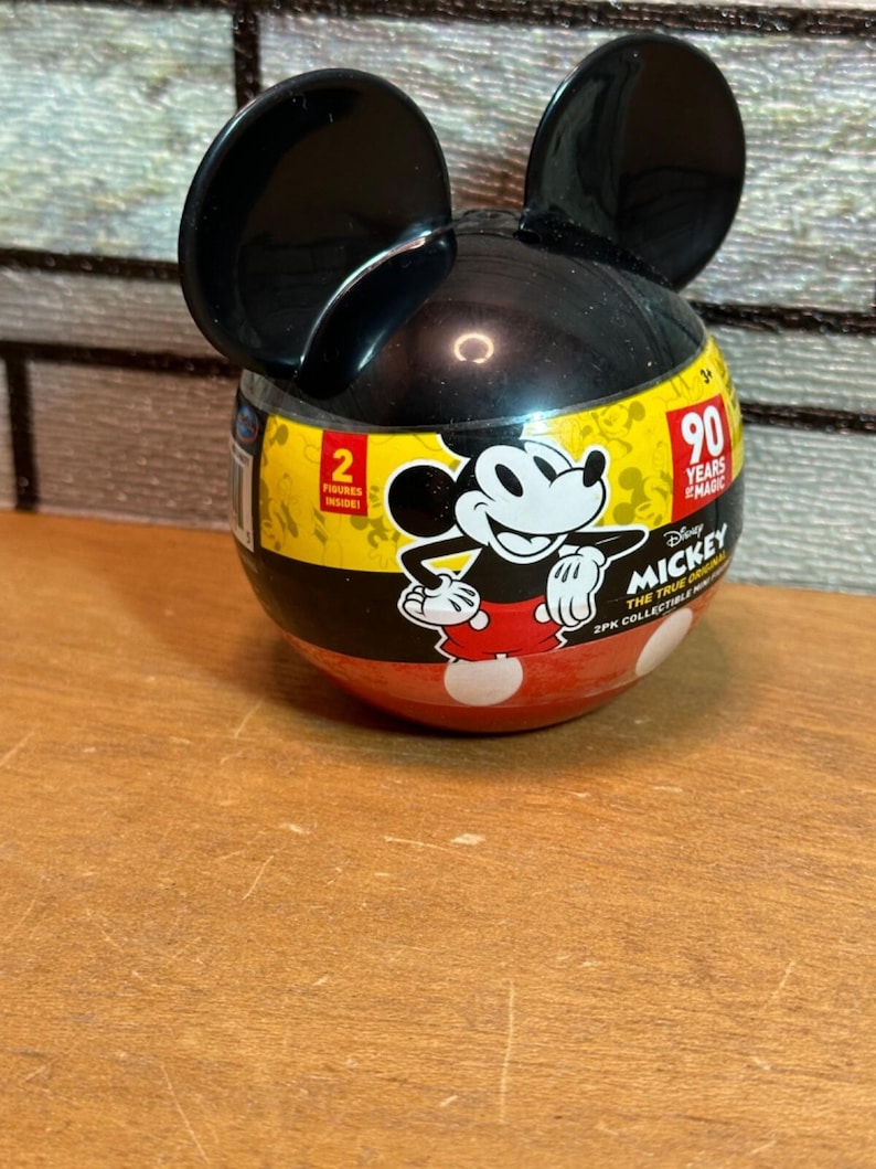May include: A Mickey Mouse-shaped container with black ears and a black and yellow body. The container features a Mickey Mouse graphic and the text "90 Years of Magic". The bottom half is red with white polka dots.