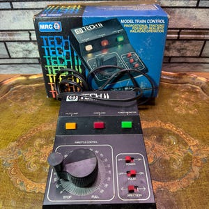 May include: A black Tech II Railmaster model train control unit with a box. The unit features a throttle control knob, coloured indicator lights, and switches for power, pulse, and direction. The box is blue and black with the text "Model Train Control".