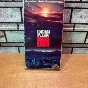 Könnte beinhalten: Eine VHS-Kassette des Films Red Dawn mit einem Sonnenuntergangshimmel, auf dem Fallschirmspringer über einer dunklen Berglandschaft absteigen. Das Cover zeigt den Titel in kyrillischer und englischer Schrift sowie MGM/UA Home Video Logos.