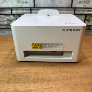 Op de afbeelding: Een witte VuPoint Photo Cube printer met een papieruitvoersleuf. De printer heeft een klein platform bovenop en een sticker met instructies. De printer staat op een houten oppervlak. De sticker luidt "PLEASE NOTE!!".