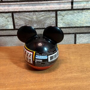 May include: A black Mickey Mouse-shaped container with two large, rounded ears. The container has a red and yellow band around the middle with small Mickey Mouse figures. The container is on a wooden surface.