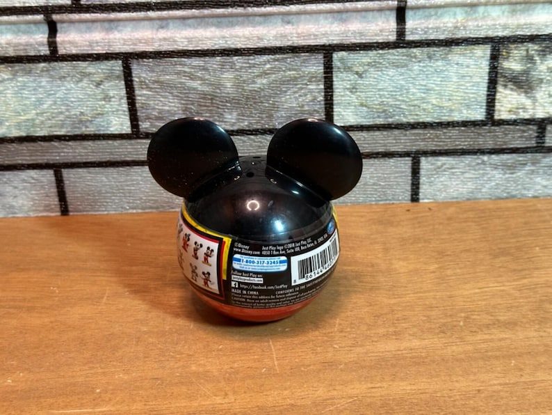 May include: A black Mickey Mouse-shaped container with two large, rounded ears. The container has a red and yellow band around the middle with small Mickey Mouse figures. The container is on a wooden surface.