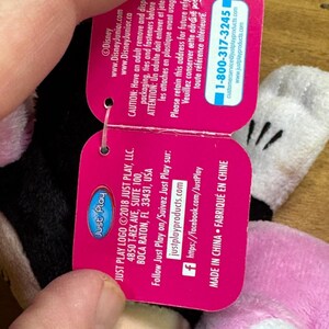 Puede incluir: Una etiqueta rosa de Just Play con el logotipo de Disney Junior, con informaci&oacute;n de contacto y una advertencia. La etiqueta est&aacute; sujeta por la mano de una persona, con un vistazo a un peluche en el fondo.