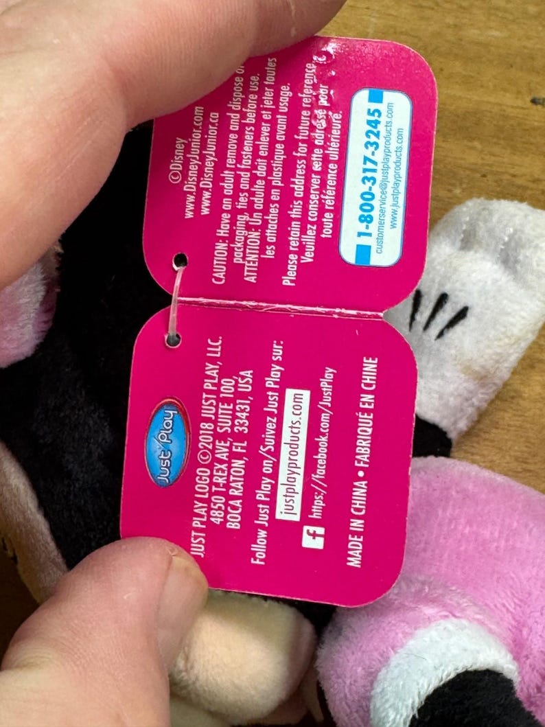 Puede incluir: Una etiqueta rosa de Just Play con el logotipo de Disney Junior, con informaci&oacute;n de contacto y una advertencia. La etiqueta est&aacute; sujeta por la mano de una persona, con un vistazo a un peluche en el fondo.