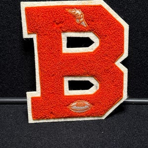 以下が含まれることがあります： 白いアウトラインとステッチが付いたオレンジ色のフェルトの文字B。文字には小さな白いフットボールと白い翼のデザインがあります。