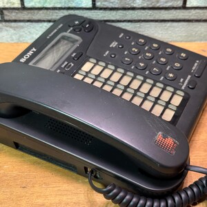 Puede incluir: Tel&eacute;fono con cable Sony negro con auricular y base. La base tiene un teclado, botones de funci&oacute;n y una peque&ntilde;a pantalla. El auricular est&aacute; conectado a la base con un cable en espiral. El tel&eacute;fono est&aacute; sobre una superficie de madera.