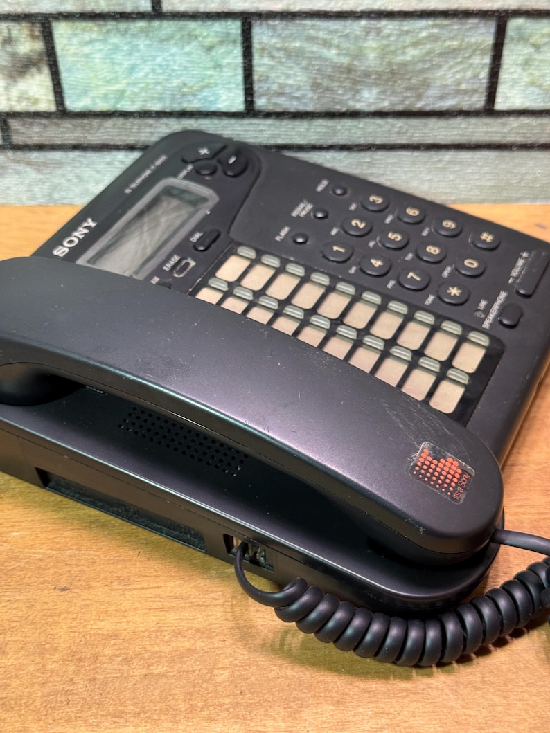 Puede incluir: Tel&eacute;fono con cable Sony negro con auricular y base. La base tiene un teclado, botones de funci&oacute;n y una peque&ntilde;a pantalla. El auricular est&aacute; conectado a la base con un cable en espiral. El tel&eacute;fono est&aacute; sobre una superficie de madera.