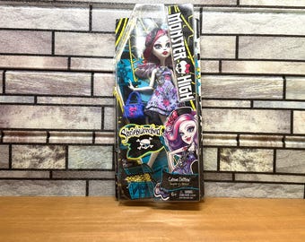 Peut inclure: Une poupée Monster High Shriek Wrecked Catrine DeMew dans son emballage d'origine. La poupée a des cheveux roses et blancs, une robe à fleurs violette et un sac à main bleu. La boîte présente le logo Monster High, un crâne et un coffre.