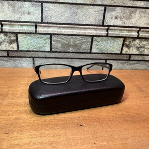 Puede incluir: Gafas con montura negra sobre un estuche rectangular negro para gafas. Las gafas tienen una forma rectangular cl&aacute;sica. El estuche est&aacute; cerrado y descansa sobre una superficie de madera, con un fondo de ladrillo.