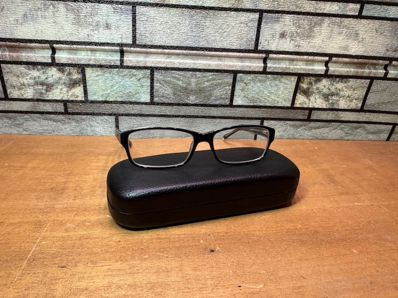 Puede incluir: Gafas con montura negra sobre un estuche rectangular negro para gafas. Las gafas tienen una forma rectangular cl&aacute;sica. El estuche est&aacute; cerrado y descansa sobre una superficie de madera, con un fondo de ladrillo.