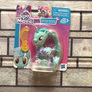 May include: A Little Pony Movie toy figure of a teal unicorn with a long, flowing mane and tail, packaged on a pink and blue card. Includes a yellow comb accessory. The packaging has the text "PONY MOVIE" and a warning about small parts.
