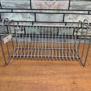 Puede incluir: Un escurreplatos de metal plateado con un diseño rectangular. El estante tiene divisores verticales para sujetar los platos y una etiqueta adjunta en el lateral. El estante está sobre una superficie de madera.