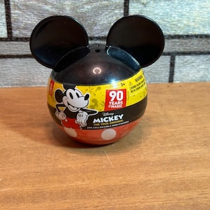 May include: A Mickey Mouse-themed collectible container with black ears. The container is black, yellow, and red, featuring Mickey Mouse graphics and the text "90 Years of Magic." The lower section is red with white polka dots.