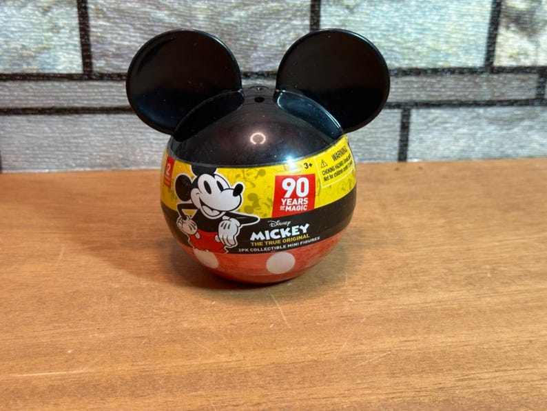 May include: A Mickey Mouse-themed collectible container with black ears. The container is black, yellow, and red, featuring Mickey Mouse graphics and the text "90 Years of Magic." The lower section is red with white polka dots.