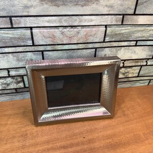 May include: A rectangular silver picture frame with a textured surface. The frame has a dark interior and is sitting on a wooden surface.