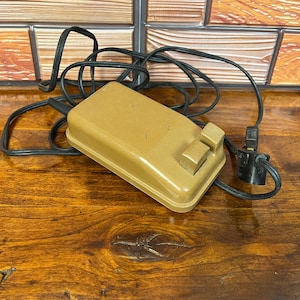 Peut inclure: Une pédale de commande de machine à coudre rectangulaire de couleur beige avec un boîtier en plastique lisse et une plaque de pression texturée. L'appareil comprend un long cordon électrique noir et une prise noire à deux broches.