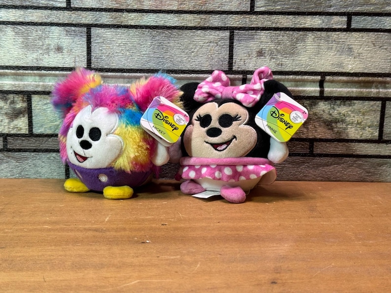 May include: Two Disney plush toys: Mickey Mouse with rainbow fur and Minnie Mouse with a pink polka dot skirt. Both have Disney tags attached. The toys are on a wooden surface with a brick background.