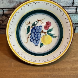Puede incluir: Un tazón de cerámica vintage con un borde amarillo y un borde verde y blanco. El tazón presenta un diseño pintado de uvas, un melocotón y una cereza.