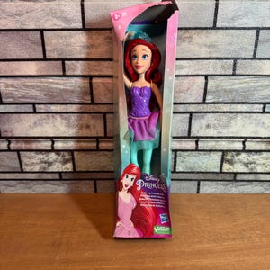 Może przedstawiać: Lalka balerina Ariel z Disney Princess w fioletowej i zielonej sukience z koroną. Lalka jest zapakowana w pudełko z r&oacute;żowym i fioletowym tłem. Na pudełku widnieją słowa "Disney Princess" i "Ballerina Princess Ariel".