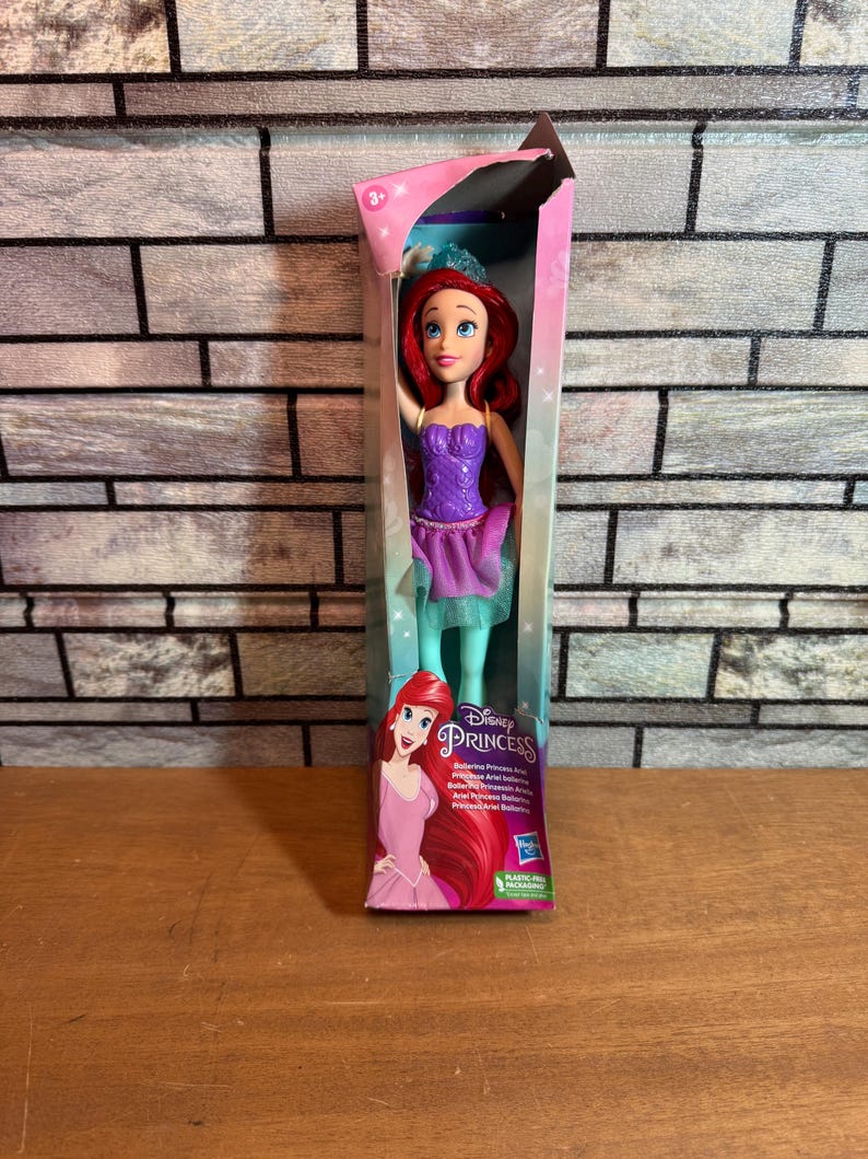 Może przedstawiać: Lalka balerina Ariel z Disney Princess w fioletowej i zielonej sukience z koroną. Lalka jest zapakowana w pudełko z r&oacute;żowym i fioletowym tłem. Na pudełku widnieją słowa "Disney Princess" i "Ballerina Princess Ariel".