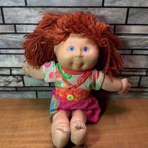 May include: A vintage doll with red yarn hair, blue eyes, and a painted face. The doll wears a floral top, a pink skirt, and has a button closure. The doll is sitting with its legs extended.
