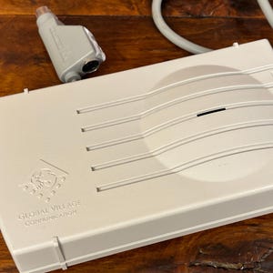 May include: A beige rectangular Global Village Communication modem with a raised curved center section featuring horizontal grooves. A gray cable with a connector plug is attached to the device, which displays the brand logo and text on the top surface.