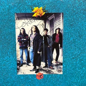 May include: A signed photograph of a band with four members. The band members are standing in front of a house with a yellow rose on the porch. The photo is framed with a blue background and a red dice is in the bottom centre.