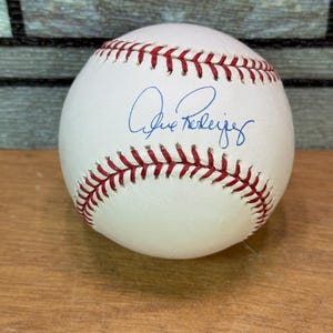May include: A white baseball with red stitching and a blue signature. The baseball is resting on a wooden surface. The stitching is visible around the entire ball.