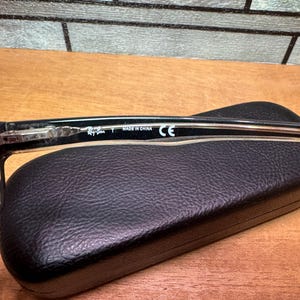 Puede incluir: Gafas Ray-Ban negras con una patilla transparente, apoyadas sobre un estuche de gafas de cuero negro. La patilla de las gafas tiene el texto "Ray-Ban T MADE IN CHINA CE". El fondo es una superficie de madera.