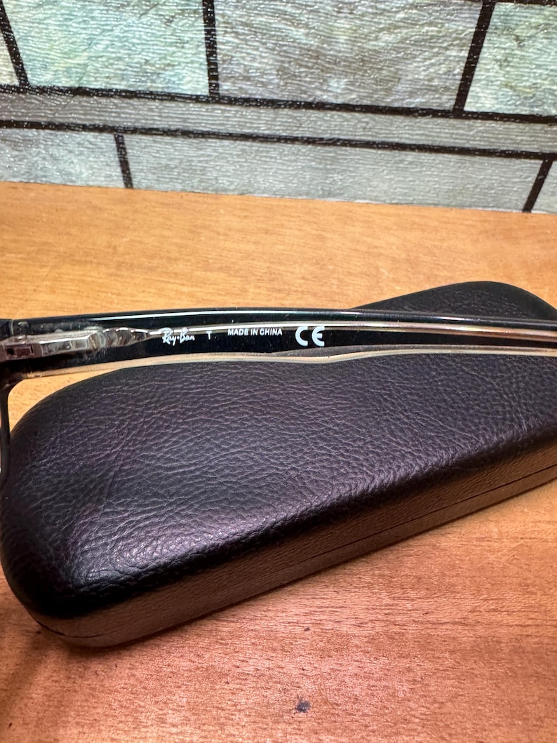 Puede incluir: Gafas Ray-Ban negras con una patilla transparente, apoyadas sobre un estuche de gafas de cuero negro. La patilla de las gafas tiene el texto "Ray-Ban T MADE IN CHINA CE". El fondo es una superficie de madera.