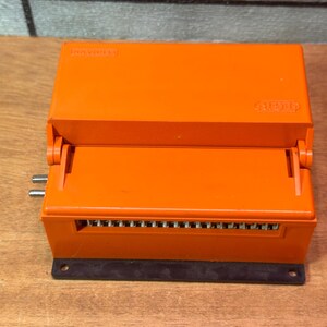 May include: An orange electrical component with a hinged lid and a black base. The component has multiple connection points and the text "Heinemann" is visible on the top. The component is on a wooden surface.