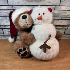 Puede incluir: Un oso de peluche marrón con un gorro de Papá Noel abraza a un muñeco de nieve. El muñeco de nieve tiene una nariz de zanahoria y un brazo de ramita. El oso tiene una nariz negra y almohadillas. El fondo es una pared de ladrillos.