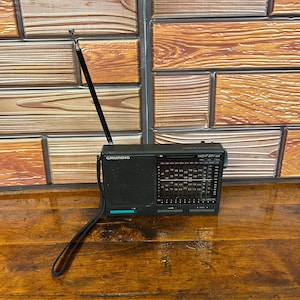 以下が含まれることがあります： 黒いGrundig Yacht Boy 220ポータブルラジオ。長方形のスピーカーグリル、周波数表示付きのチューニングダイヤル、長く伸びる金属製アンテナ、そして本体の側面に取り付けられた黒い布製のキャリングストラップが特徴です。