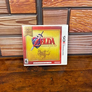 May include: A Nintendo 3DS video game case for The Legend of Zelda: Ocarina of Time 3D. The cover features a yellow background with a red border, a blue shield with a sword, and a silhouette of a rider on horseback. The Nintendo Selects logo is visible.