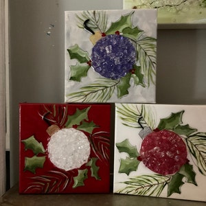 Peut inclure: Trois peintures carrées sur le thème de Noël. Chacune présente une décoration texturée en violet, blanc et rouge, entourée de houx et d'aiguilles de pin. Les fonds sont rouges, blancs et bleu clair.