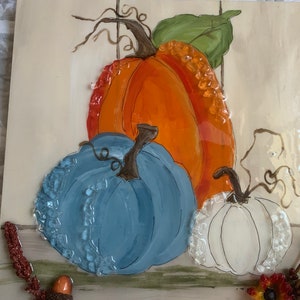May include: A painted ceramic wall decor featuring three pumpkins. The pumpkins are painted in shades of orange, blue, and white. The background is a light beige with a green leaf and brown vines.