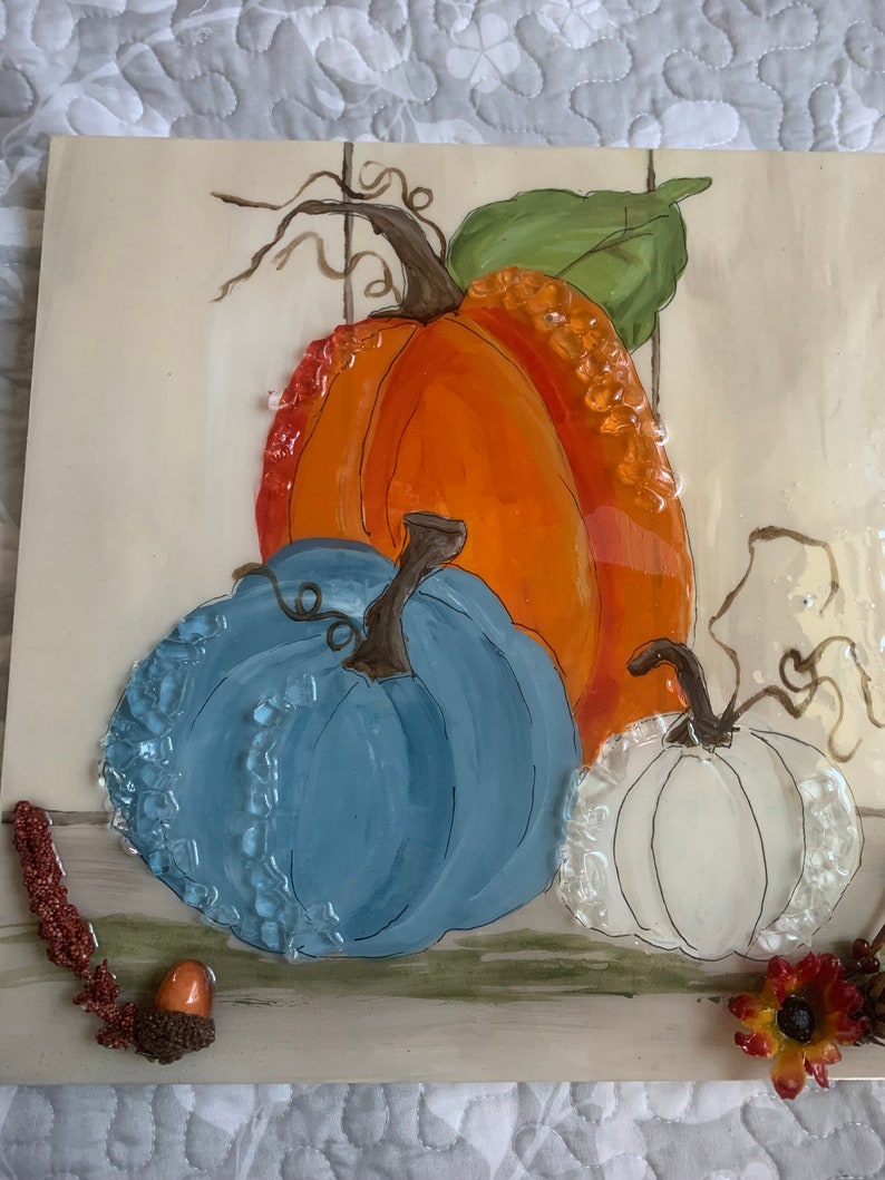 Peut inclure: Une d&eacute;coration murale en c&eacute;ramique peinte repr&eacute;sentant trois citrouilles. Les citrouilles sont peintes dans des tons d'orange, de bleu et de blanc. L'arri&egrave;re-plan est de couleur beige clair avec une feuille verte et des vignes brunes.
