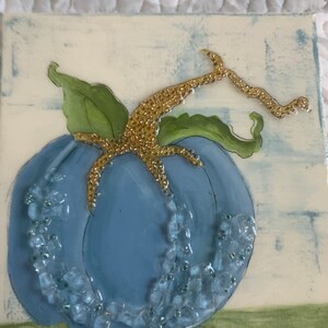 Peut inclure: Une citrouille bleue avec une tige dorée et des perles blanches et bleues autour. La citrouille est peinte sur une toile avec un fond blanc.