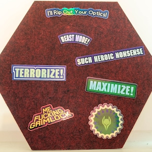 Könnte beinhalten: Eine rote sechseckige Korktafel mit verschiedenen bunten Aufklebern mit Text wie "I'll Rip Out Your Optics!" und "BEAST MODE!"