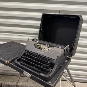 May include: A vintage Underwood typewriter with a dark gray textured finish and black circular keys. The typewriter is housed in its original black carrying case, which is open, and is mounted on a set of thin, silver-colored metal legs.