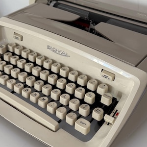 May include: Cream Royal typewriter with a silver top. The keyboard has white keys with black lettering. The word "ROYAL" is printed on the front. The typewriter has a column set and a shift key.