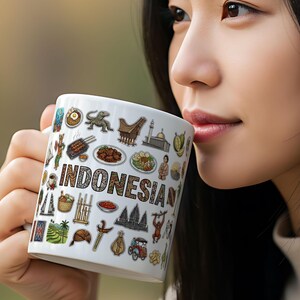 Op de afbeelding: Witte keramische mok met het woord "INDONESIAN" en diverse Indonesische culturele iconen. De mok wordt door een persoon vastgehouden. De mok toont illustraties van eten, bezienswaardigheden en traditionele items.