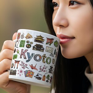 May include: White ceramic mug with a colourful design of Kyoto landmarks and the city's name. The design includes temples, cherry blossoms, and a paper crane. The mug is held by a person.