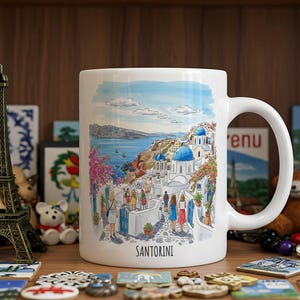 May include: White ceramic mug featuring a watercolor illustration of Santorini, Greece, with blue-domed buildings, the sea, and the word "SANTORINI". The mug has a curved handle and is surrounded by decorative items.