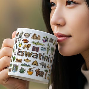 Może przedstawiać: Biały ceramiczny kubek z kolorowymi ilustracjami i napisem "Eswatini". Kubek zdobią różne obrazy, w tym lew, nosorożec i tradycyjna chata. Kubek jest trzymany przez osobę.