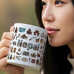 Op de afbeelding: Witte keramische mok met het woord "Nepal" en diverse kleurrijke illustraties van Nepalese bezienswaardigheden en culturele symbolen. De mok wordt door een persoon vastgehouden, de achtergrond is wazig.