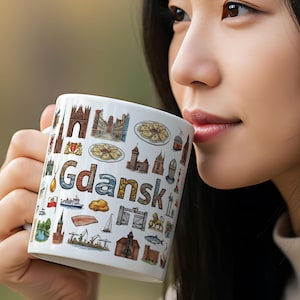 May include: White ceramic mug featuring the word "Gdansk" in colorful letters, surrounded by illustrations of landmarks and symbols. The mug has a handle and is held by a person.