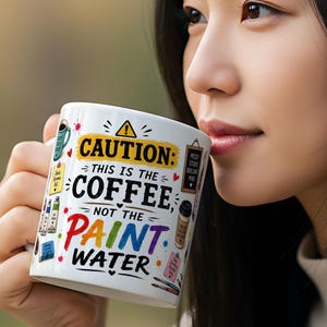 May include: White ceramic mug with the text "CAUTION: THIS IS THE COFFEE, NOT THE PAINT WATER" in black, red, and purple. The mug has colorful illustrations of paint supplies and a coffee cup. The mug is held by a person.