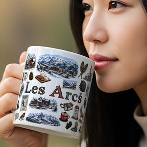 Op de afbeelding: Witte keramische mok met de tekst "Les Arcs" en diverse illustraties van bergen en gebouwen. De mok wordt tegen de mond gehouden. De mok heeft een oor.